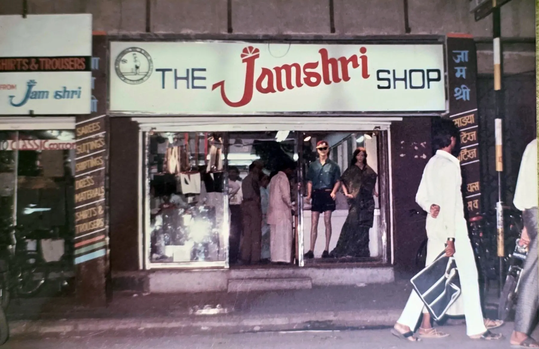 The Jamshri Shop storefront, its fascia lettered in English and Devanagari, with sarees, suitings and shirtings listed down the pillar and two dressed mannequins in the window